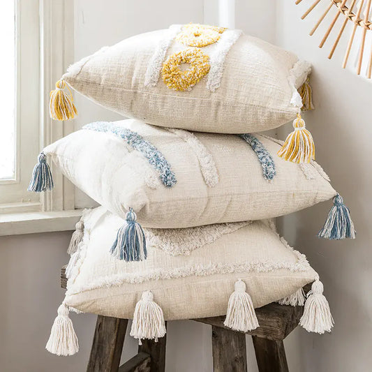 Stack of decorative pillows with tassels on a wooden stool against a neutral wall.