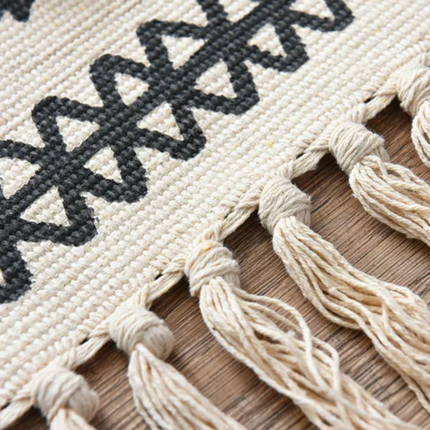 Close-up of a woven mat with black geometric patterns and white tassels on a wooden surface.