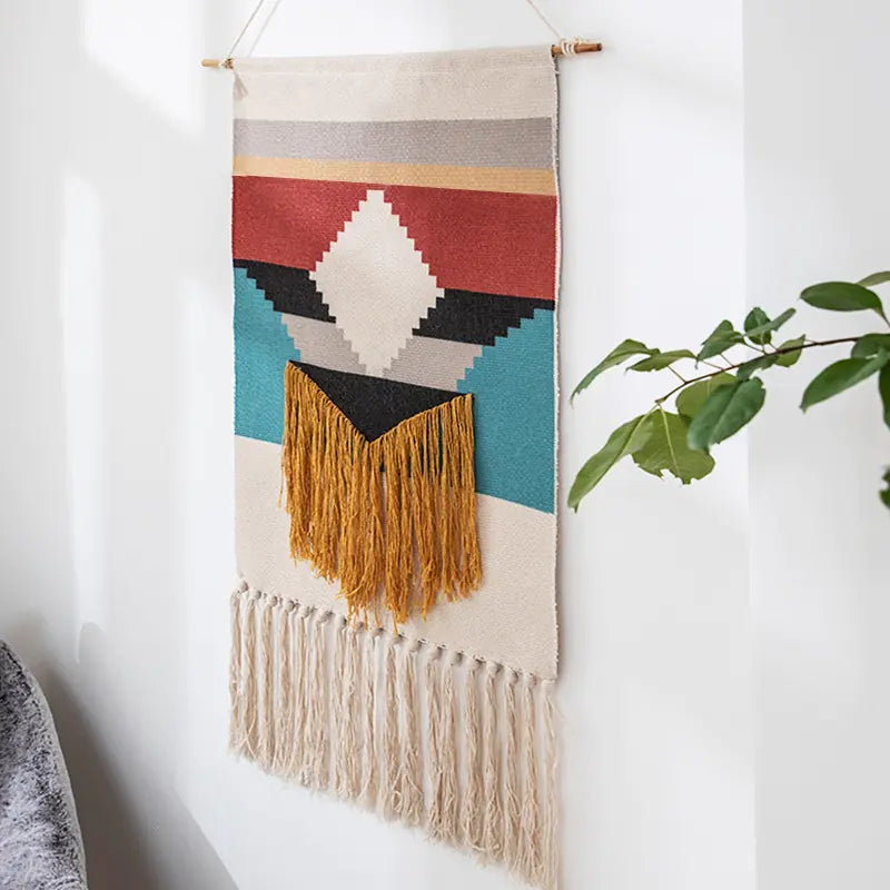 Colorful woven wall hanging with geometric patterns and fringes on a white wall.