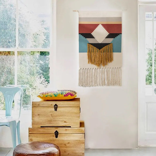 Decorative wall hanging with geometric pattern and fringes above wooden storage boxes in a room with large windows.